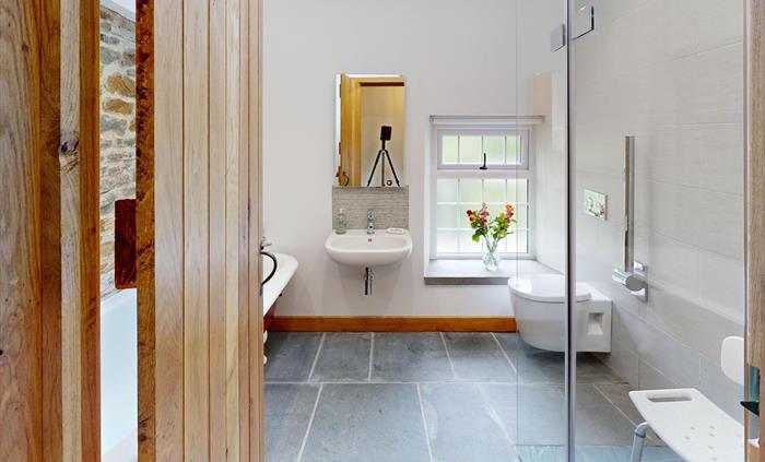 A bright bathroom with a walk-in shower, shower seat, wall-mounted toilet, sink, and a window with a vase of flowers, hand rails